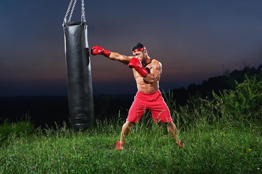 Full length shot of a young strong boxer with sexy hot ripped body practicing outdoors working out on a punching bag copyspace sunset on the background dusk epic skies ambitious concentrated fighter.