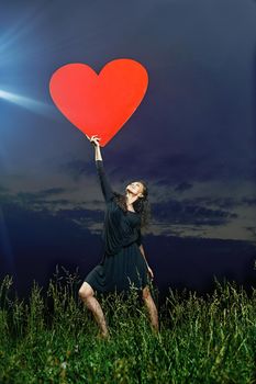 beautiful, flexible, smiling, curly dancer posing on grassy field with a red heart in his hands in front of the evening sky.