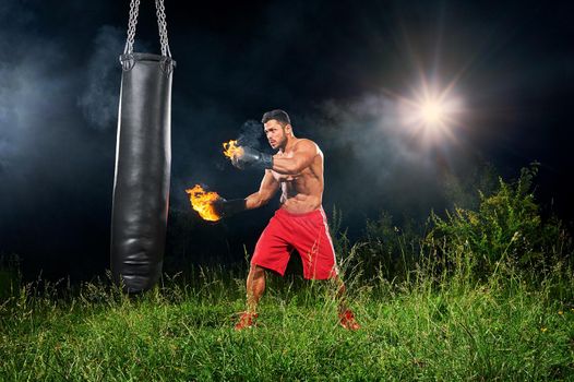 Young athletic male fighter working out with a punching bag outdoors at night with fire burning on his boxing gloves copyspace power strength masculinity fierce fiery flames hot sexy sportsman focused.