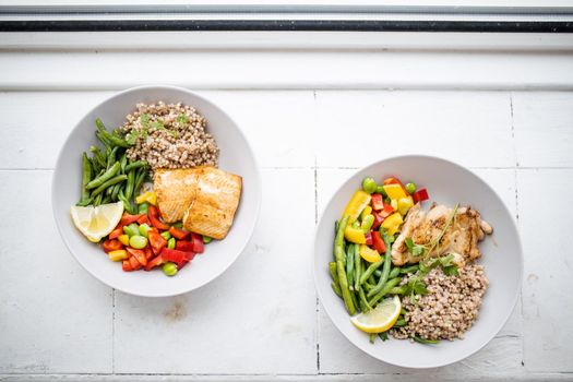 Top view of chicken and salmon dishes with buckwheat, green beans, broad beans, tomato, and pepper slices. Nutritious dishes with vegetables and meat on white plates. Healthy balanced diet