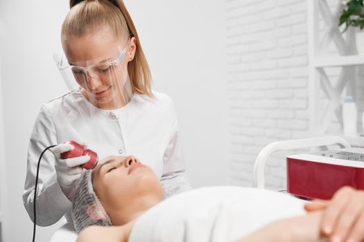 Side view of young cute beautician in protective mask doing cleaning procedure for face with special machine for young woman. Concept of beauty procedures for improvements skin condition. 