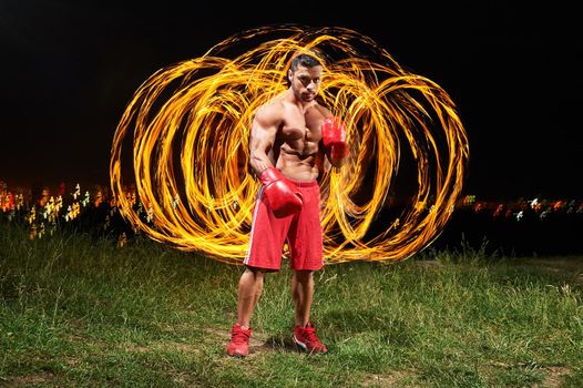 Muscular shirtless handsome sexy young man wearing boxing gloves posing forcefully outdoors at night with fire and flames behind his back copyspace sportsman sport motivation athlete winner fighting .