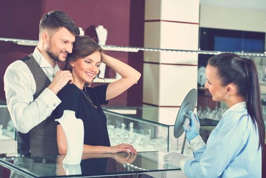 Making her happy. Beautiful happy woman trying on a necklace at the jewelry store with her handsome loving boyfriend and professional jeweler helping luxury sale discount gift present love concept