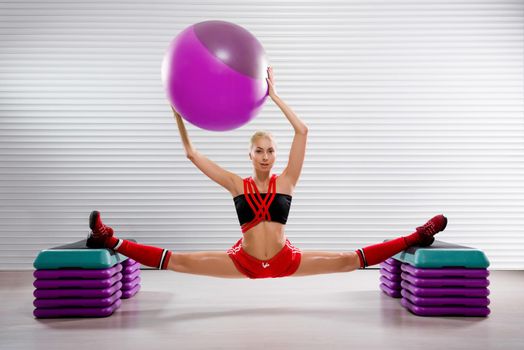 Stunning flexible female gymnast holding a fit ball over her head while practicing splits at the fitness studio flexibility stretching exercise workout trainer body strength motivation sports concept.