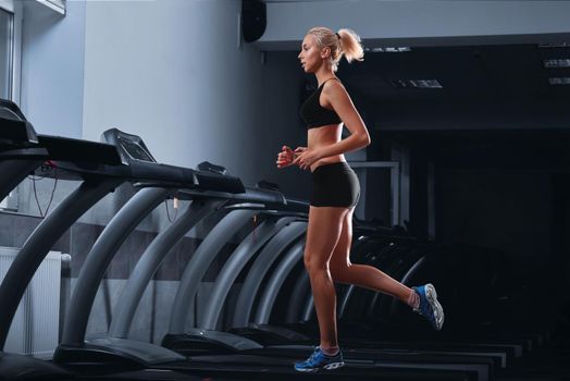 Horizontal full length shot of an attractive fit young woman with perfect strong muscular body wearing sports top and shorts jogging on a treadmill at the health club diet slimming toning motivation .