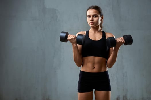 Isolated portrait of pumped fitnesswoman in black sportswear posing on gray studio background, loft interior. Front view of muscular caucasian woman training with dumbbells and looking at camera.