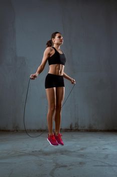 Side view of pretty female bodybuilder in black sport suit with perfect body using skipping rope in gym. Young athletic brunette woman jumping in hall, loft gray atmosphere. Concept of sport.