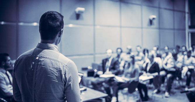 Speaker giving a talk on corporate business conference. Unrecognizable people in audience at conference hall. Business and Entrepreneurship event. Blue toned black and white.