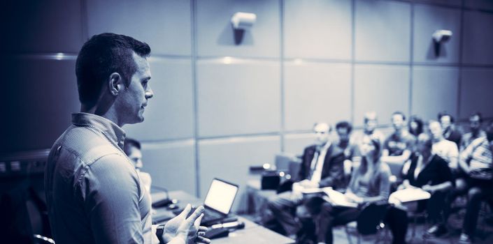 Speaker giving a talk on corporate business conference. Unrecognizable people in audience at conference hall. Business and Entrepreneurship event. Blue toned black and white.