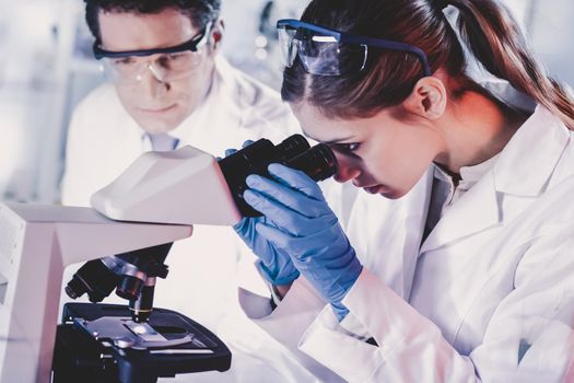 Life scientists researching in laboratory. Attractive female young scientist and her doctoral supervisor microscoping in their working environment. Healthcare and biotechnology.