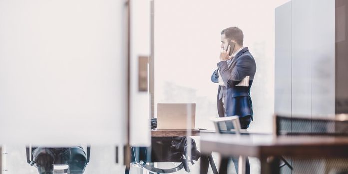 Businessman talking on a mobile phone while looking through modern corporate office window, holding financial newspaper.