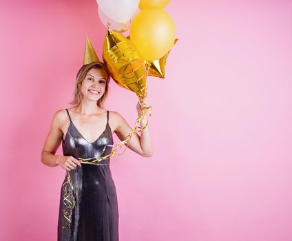 Birthday party. Young blond smiling woman wearing birthday hat holding golden balloons celebrating birthday on pink background with copy space