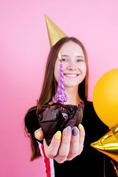 Birthday party. Teenager girl in golden birthday holding cake with candle, making a wish over pink background. Focus on foreground