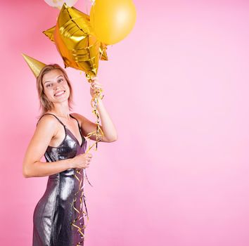 Birthday party. Young blond smiling woman wearing birthday hat holding golden balloons celebrating birthday on pink background with copy space