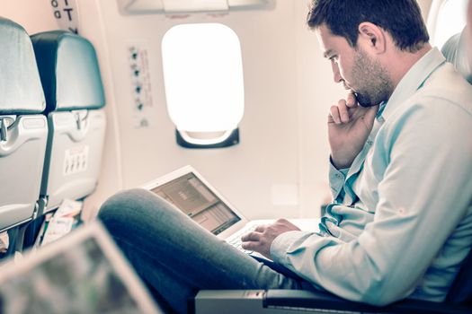 Casually dressed middle aged man working on laptop in aircraft cabin during his business travel. Shallow depth of field photo with focus on businessman eye.