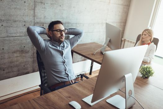Young handsome stressed businessman sitting at the office and pensive looking at computer with arms behind head.