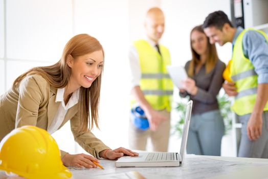 Smiling young woman constructor at the laptop in the office. Her team is at the background. Selective focus.