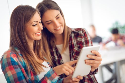 Two young businesswomen holding digital tablet and smiling, one woman pointing. 