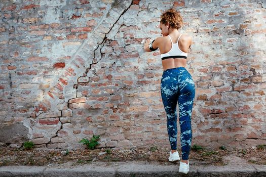 Rear view of a young muscular woman standing up against the wall and resting after jogging.