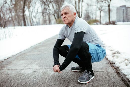 Active senior making pause crouching, during stretching and doing exercises in public park during the winter training outside in. Copy space.