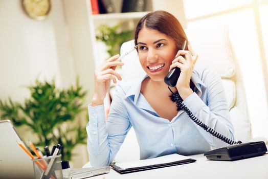 Successful young woman working at her desk in the office. She is using phone and thinking. 