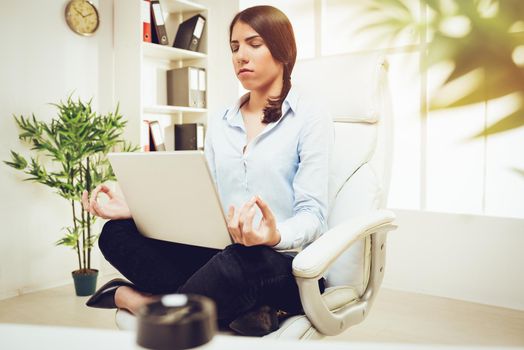 Beautiful young businesswoman sitting in lotus pose while meditating in the office. She is practicing yoga and holding a laptop in her lap.