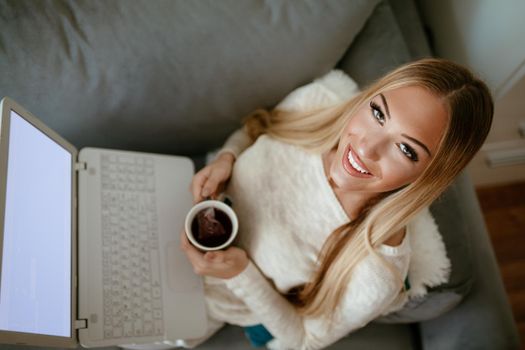 Attractive smiling girl sitting at her living room with cup of tea using laptop. She is surfing on the net and checking social media. Looking at camera. Top view.