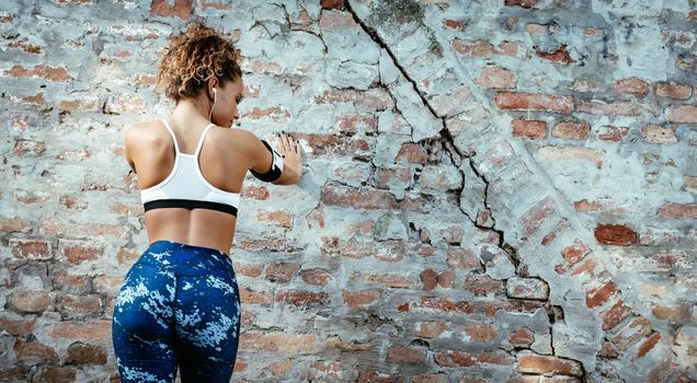 Rear view of a young muscular woman standing up against the wall and resting after running.