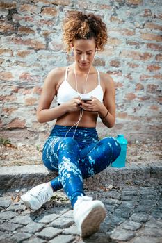 Young sporty woman with headphones taking a break after training in nature and using mobile phone.