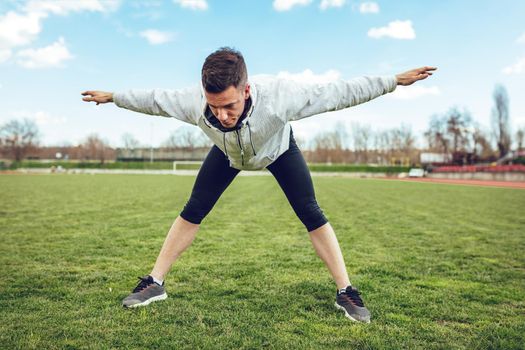 Active young man stretching and doing exercises in the public place on empty stadium. 
