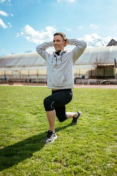 Active young man stretching and doing one leg crouching exercises in the public place on empty stadium. 