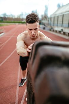 Close-up of young muscular man flipping a huge tire for training muscles at the tartan track on the stadium.