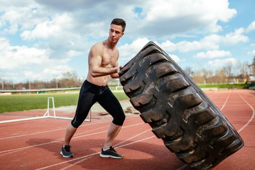 Young muscular man lifting up a huge tire for training muscles at the tartan track on the stadium.
