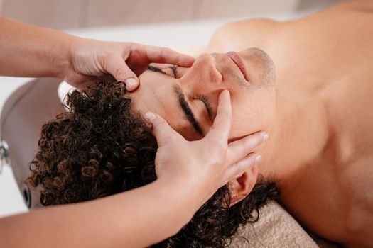 Close-up of a handsome healthy young man enjoying relaxing facial massage at beauty salon.