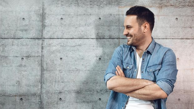 Young successful smiling entrepreneur standing in front of a concrete wall with crossed hands.