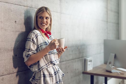 Young beautiful successful female entrepreneur standing in the office,l with cup of coffee in her hand.