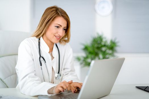 Young female doctor in her consulting room,  writing on laptop.