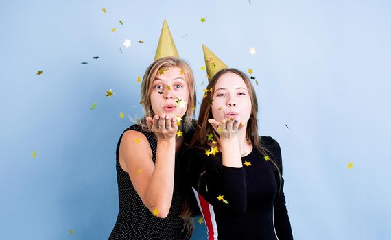 Birthday party. Two young women in birthday hats holding balloons celebrating birthday over blue background, blowing confetti