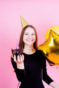 Birthday party. Teenager girl in golden birthday holding muffin with a candles, making a wish over pink background