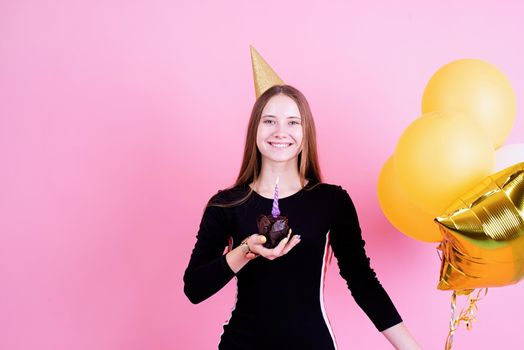 Birthday party. Teenager girl in golden birthday holding muffin with a candles, making a wish over pink background