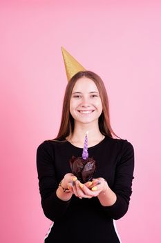 Birthday party. Teenager girl in golden birthday holding muffin with a candles, making a wish over pink background