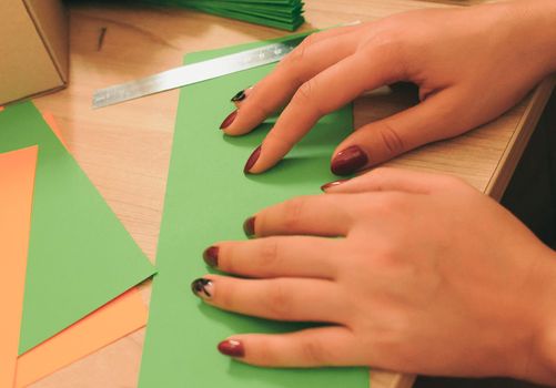 In the foreground, a girl prepares blanks for an origami fan, sheets of colored paper, scissors on a wooden table. Several blanks for a fan. Template for design, advertising or text.
