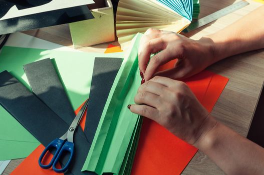 In the foreground, female hands are preparing blanks for an origami fan, sheets of colored paper, scissors on a wooden table. Several blanks for a fan. Template for design, advertising or text.