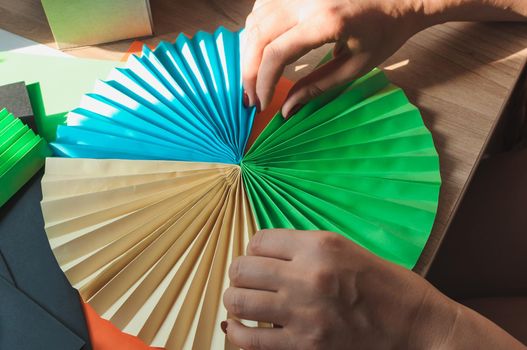 In the foreground female hands glue origami fans, sheets of colored paper, scissors on a wooden table. Several blanks for the fan. Template for design, advertising or text.