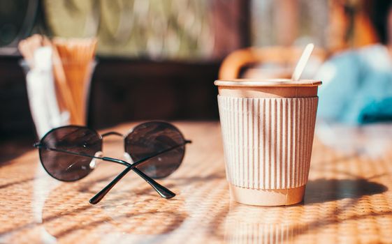 Disposable paper coffee cup with holder on table in cafe with wooden stirrer,near sunglasses.Environmentally friendly lifestyle.Zero Waste,Save The Planet,Earth Day,No Plastic,Recycling Concept