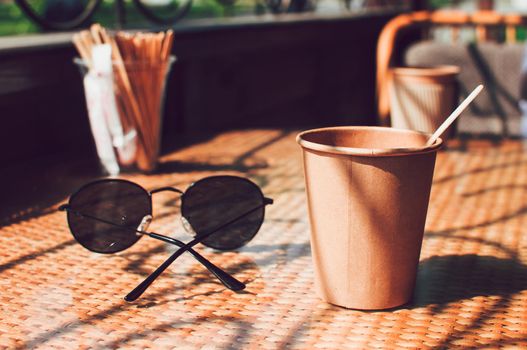 Disposable paper coffee cup on table in cafe with wooden stirrer,near sunglasses.Environmentally friendly lifestyle.Zero Waste,Save The Planet,Earth Day,No Plastic,Recycling Concept