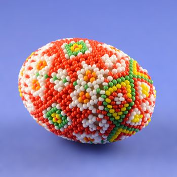 Closeup of some beautiful bead work designed over a chicken egg and placed on its side while over a blue background.