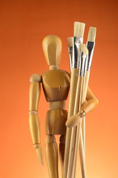 An Artists wooden Dummy Mannequin stands with some dry paintbrushes over an orange background.