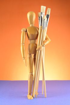 A full body view of an Artists Dummy Mannequin standing and holding various sized dry paintbrushes over a blue and orange background.