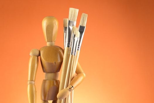 An Artists wooden Dummy Mannequin stands with some dry paintbrushes over an orange background.